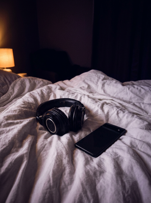 A dimly lit bedroom at night with over-ear headphones and a phone resting on a rumpled white duvet, a warm bedside lamp glowing in the corner.