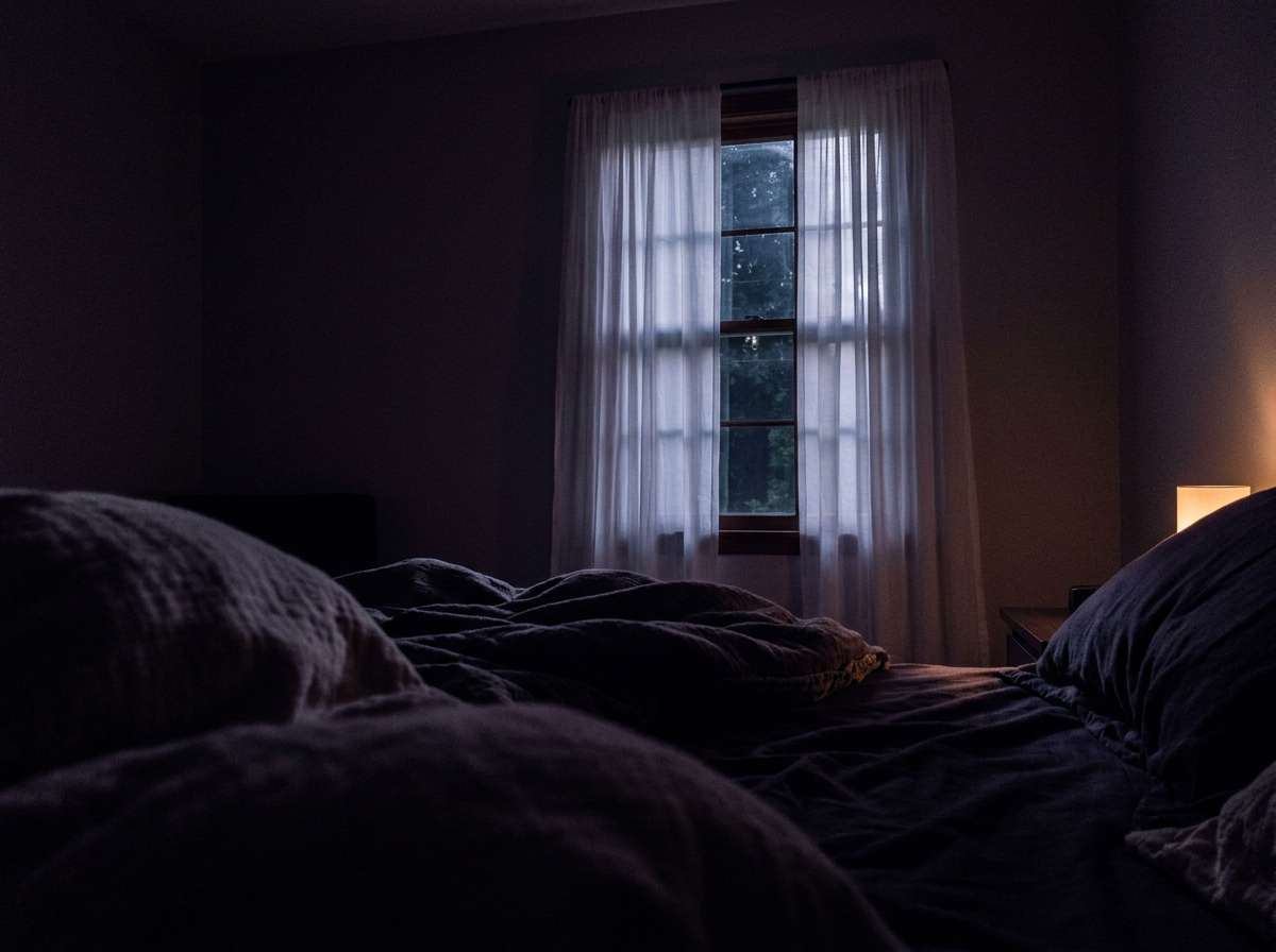 A dark bedroom at night, a window with sheer curtains letting in faint moonlight and a small warm lamp glowing beside the bed.